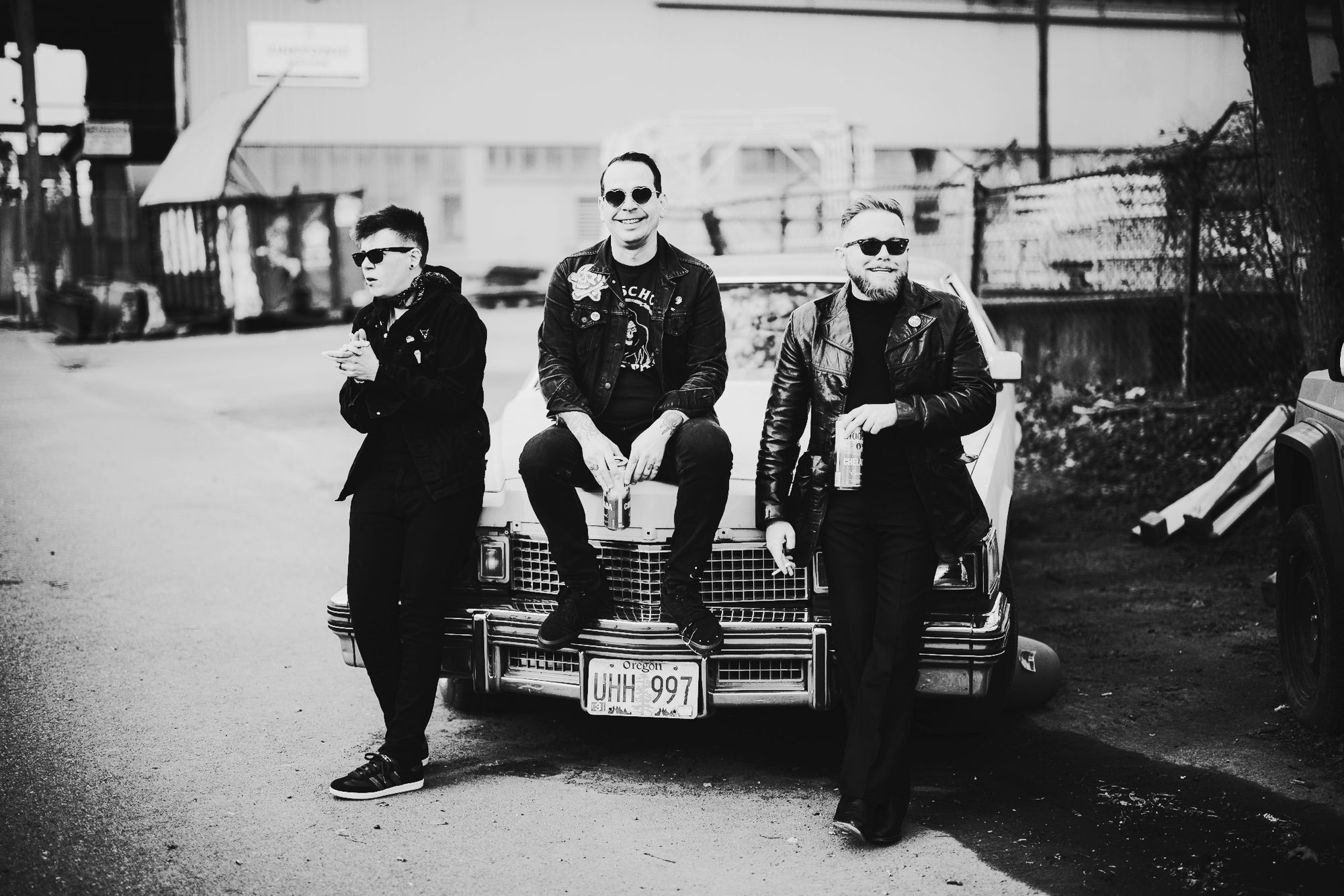 Tragic Lovers in a gravel lot, leaning on and sitting on the hood of a white Cadillac.